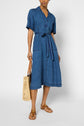 A person wearing a belted denim midi dress with short sleeves, tan sandals, and holding a woven beige tote bag, standing against a plain light background.