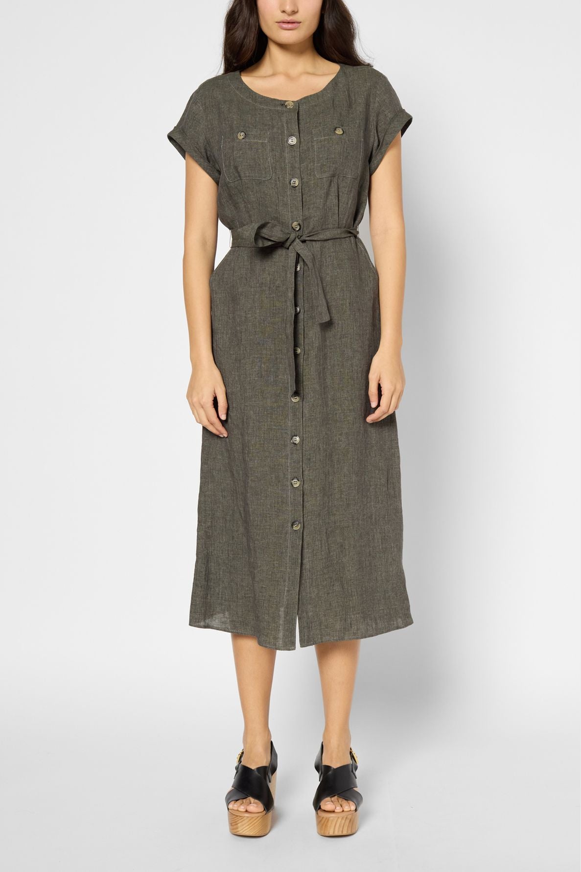Woman wearing a short-sleeved, button-front, olive green midi dress with a waist tie, standing against a plain background. She has black platform sandals and is facing forward with arms by her sides.