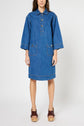 A woman wearing a loose-fitting, knee-length denim dress with large front pockets, button details, and wide sleeves. She stands facing forward and wears brown platform sandals.