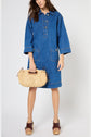Woman wearing a loose blue denim dress, holding a woven beige handbag, and brown platform sandals. Her hair is loose and her face is partially out of frame. She stands against a plain white background.
