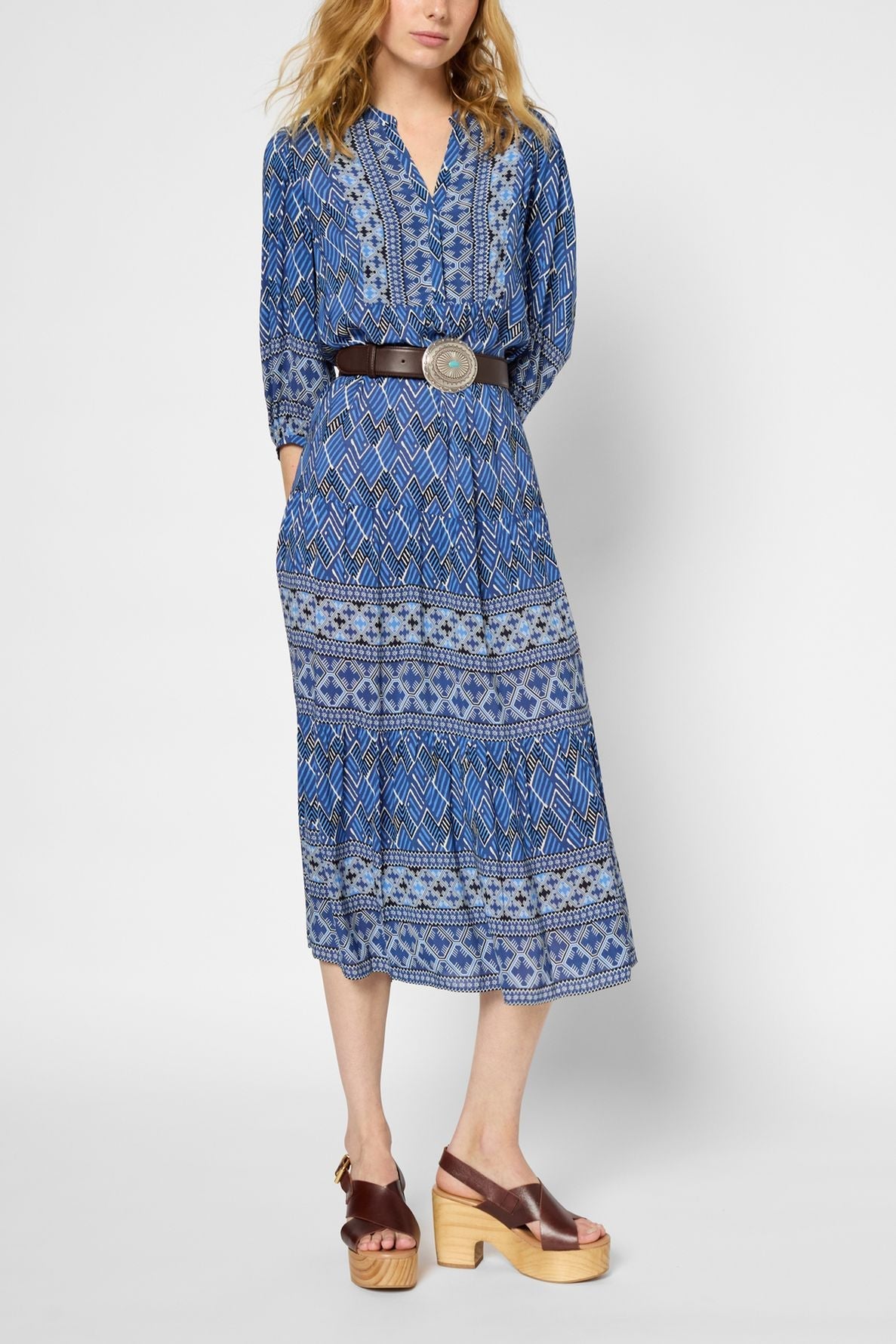 A woman wearing a patterned blue midi dress with a wide belt and brown platform sandals stands against a plain background. Her hands are in her dress pockets, and her blonde hair is loose.