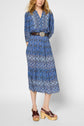 A woman wearing a patterned blue midi dress with a wide belt and brown platform sandals stands against a plain background. Her hands are in her dress pockets, and her blonde hair is loose.