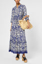 A person wearing a robe longue imprimée bleu et blanc au motif floral, des sandales noires à plateforme, et tenant un sac à main en paille avec des poignées en bois, se tient debout contre un fond uni.