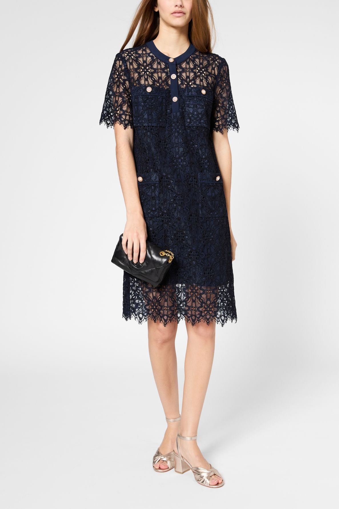 Woman wearing a short-sleeved navy lace dress, holding a black clutch, and wearing silver sandals. Her head is not fully visible in the image.