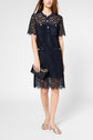 Woman wearing a short-sleeved navy lace dress, holding a black clutch, and wearing silver sandals. Her head is not fully visible in the image.