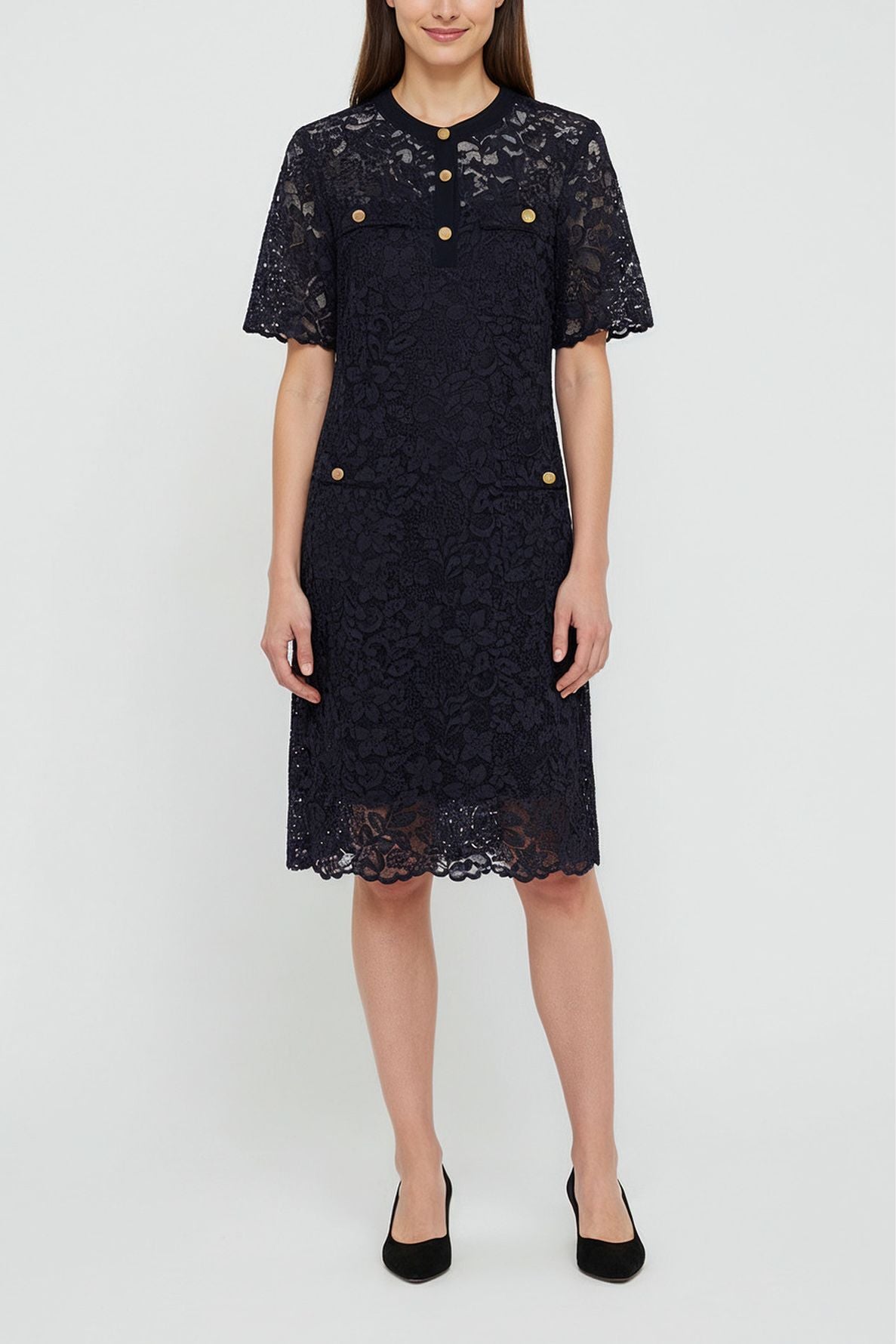 A woman wears a knee-length, short-sleeved, navy lace dress with gold buttons on the chest and pockets. She stands facing forward, with her arms relaxed by her sides, and wears black shoes.