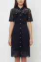 Woman wearing a knee-length, short-sleeved, navy lace dress with buttons down the front, two chest pockets, and a col chemise neckline.