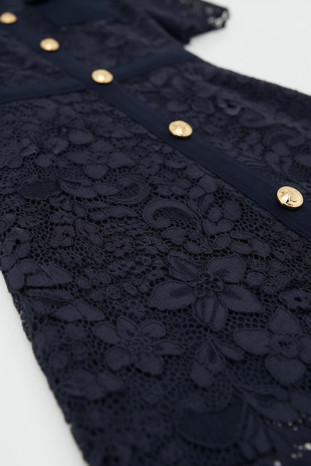 Close-up of a navy blue robe en dentelle featuring floral patterns and a row of gold buttons down the front. The intricate lace detail is prominent, giving this elegant robe longue with col chemise a refined and textured appearance.