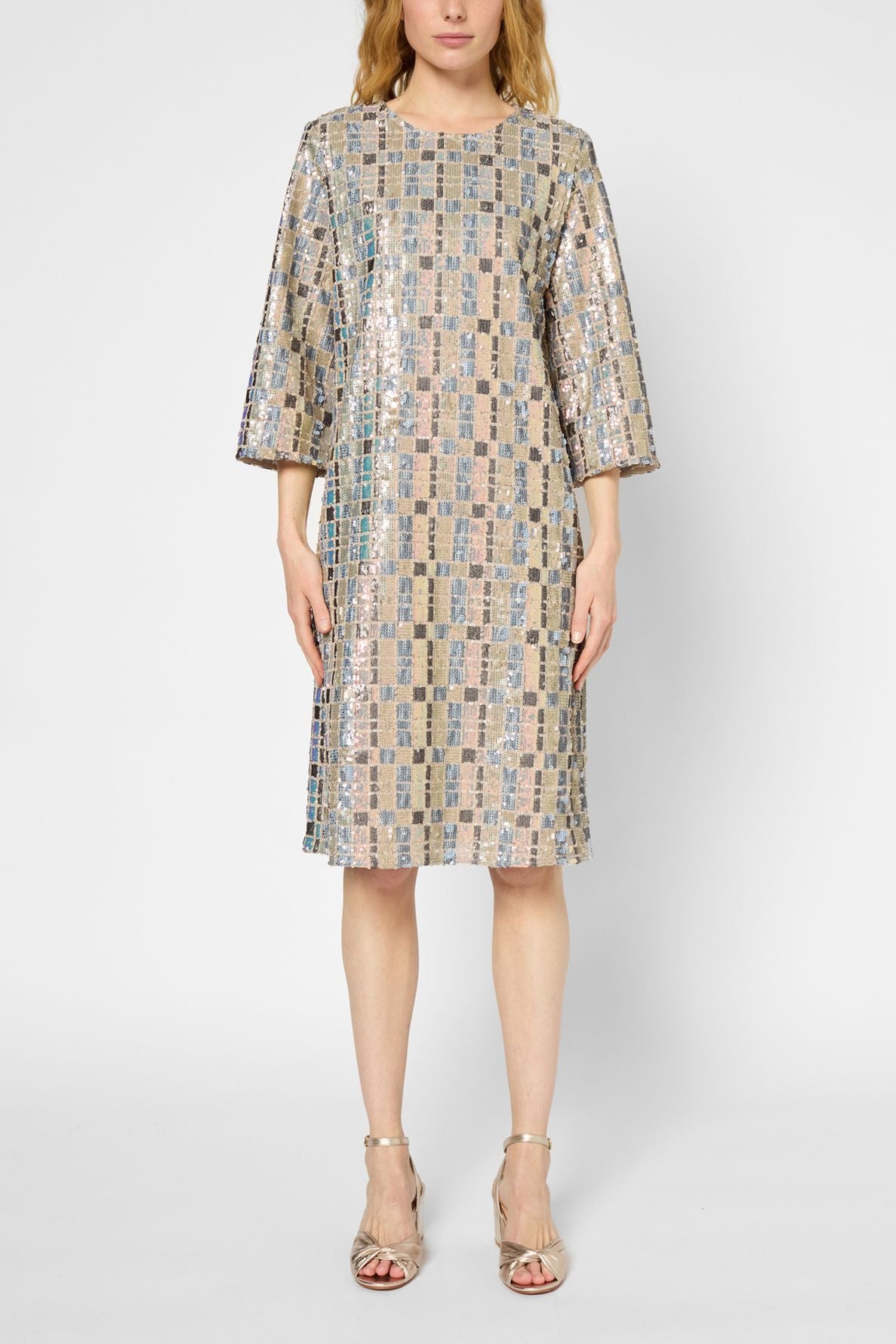 Woman wearing a knee-length, loose-fitting robe droite sequins with a geometric sequin pattern, three-quarter sleeves, and metallic open-toe heels, standing against a plain white background.