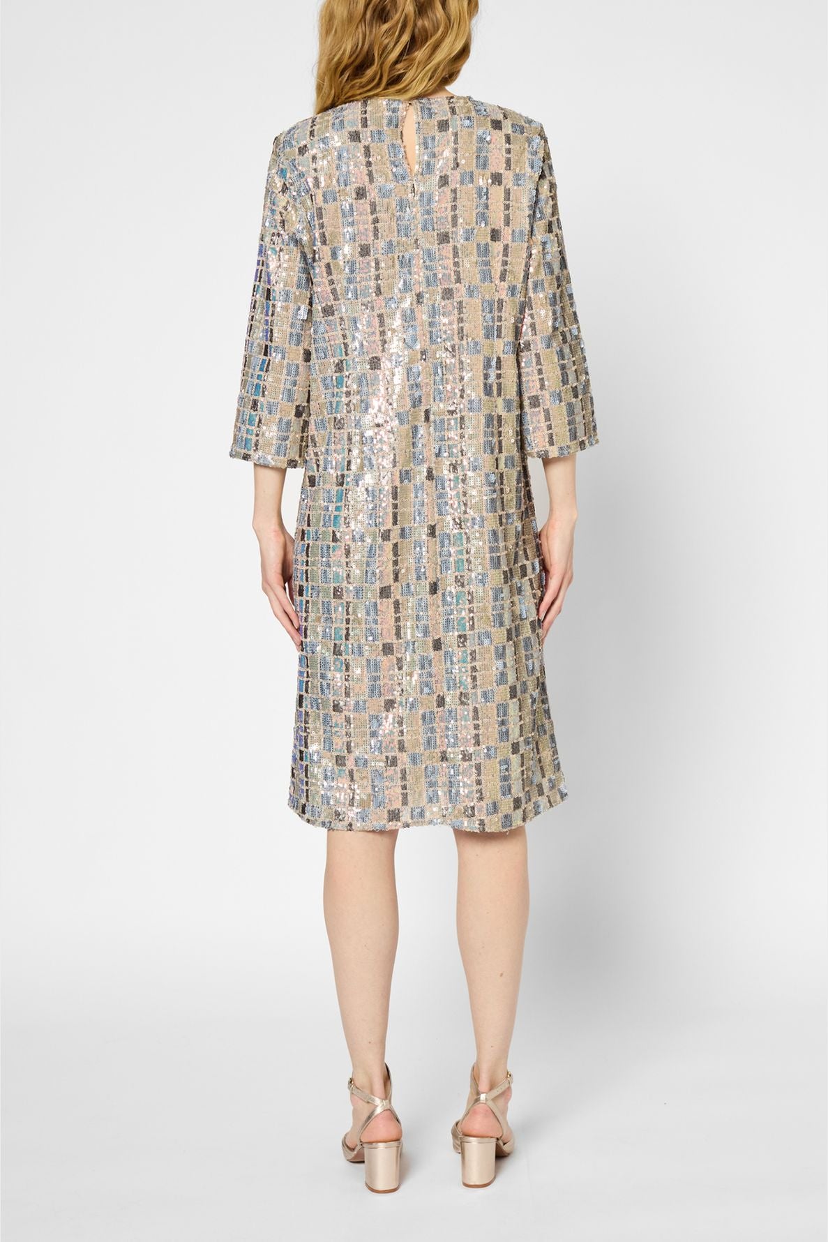A person wearing a knee-length, long-sleeved robe droite sequins with a shiny checked pattern stands facing away, showing the back of the dress. They have light-colored heels and long, wavy blonde hair.