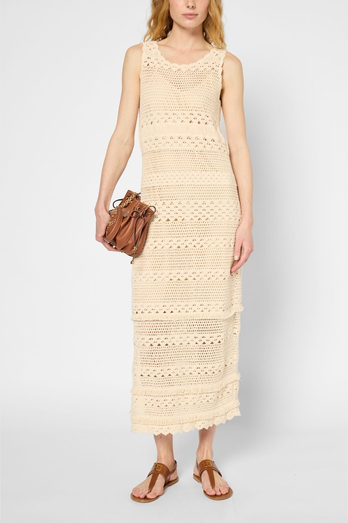 A woman wearing a sleeveless, cream-colored crochet maxi dress, brown sandals, and holding a small brown handbag stands against a plain white background.