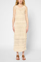 A woman wearing a sleeveless, ankle-length, cream-colored crochet dress and brown sandals stands against a plain light gray background.