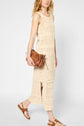 A person wearing a sleeveless, cream-colored crochet dress, brown sandals, and carrying a small brown handbag walks against a plain white background.
