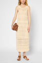 A woman in a sleeveless, beige crochet dress stands holding a small brown purse. She wears flat brown sandals and has wavy, blonde hair.