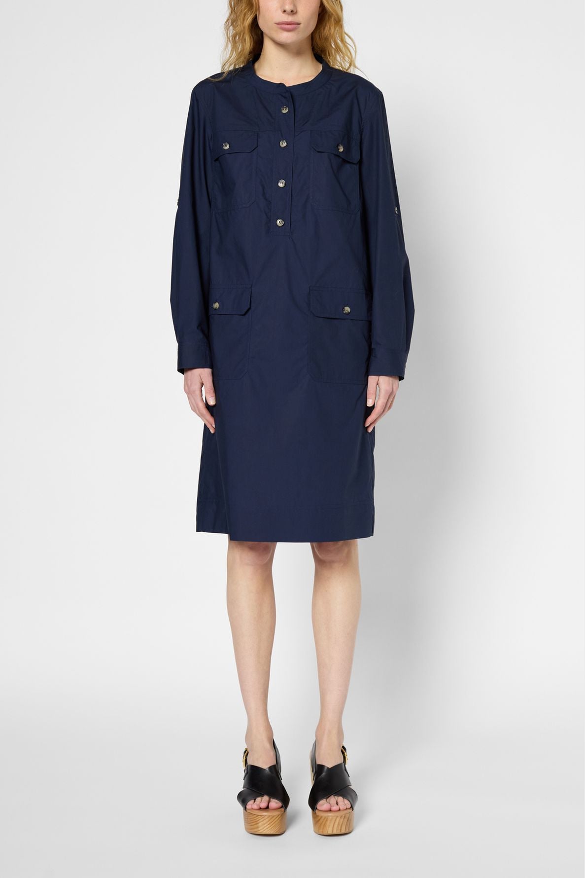 A person wearing a navy blue, long-sleeve, knee-length dress with front pockets and black platform sandals stands against a plain light background.