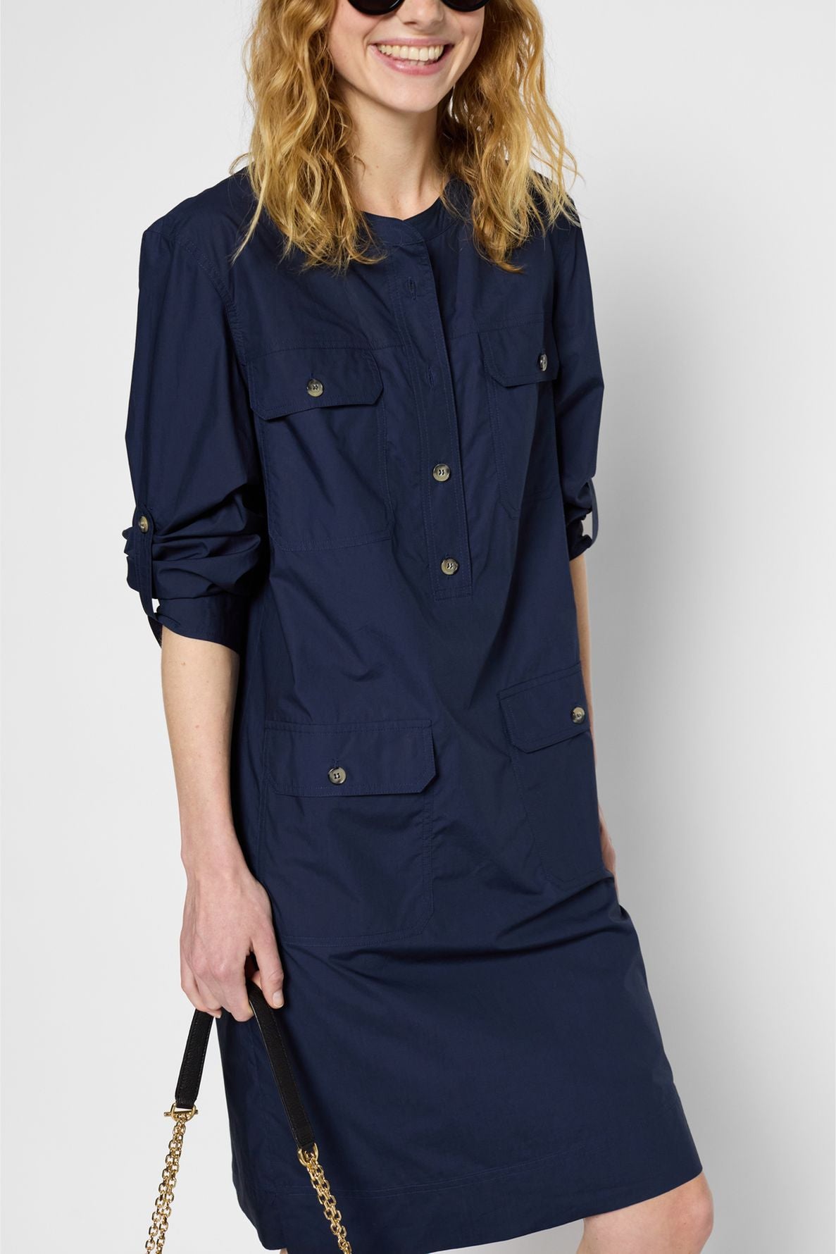 Woman smiling, wearing a navy blue long-sleeve shirt dress with buttons and front pockets. She holds a black purse with a gold chain. Her sleeves are rolled up and her wavy hair is loose.