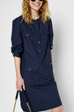 Woman smiling, wearing a navy blue long-sleeve shirt dress with buttons and front pockets. She holds a black purse with a gold chain. Her sleeves are rolled up and her wavy hair is loose.