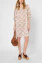 A woman wearing a patterned knee-length dress with 3/4 sleeves holds a brown handbag and wears brown platform sandals. She has loose, wavy hair and stands against a plain white background.
