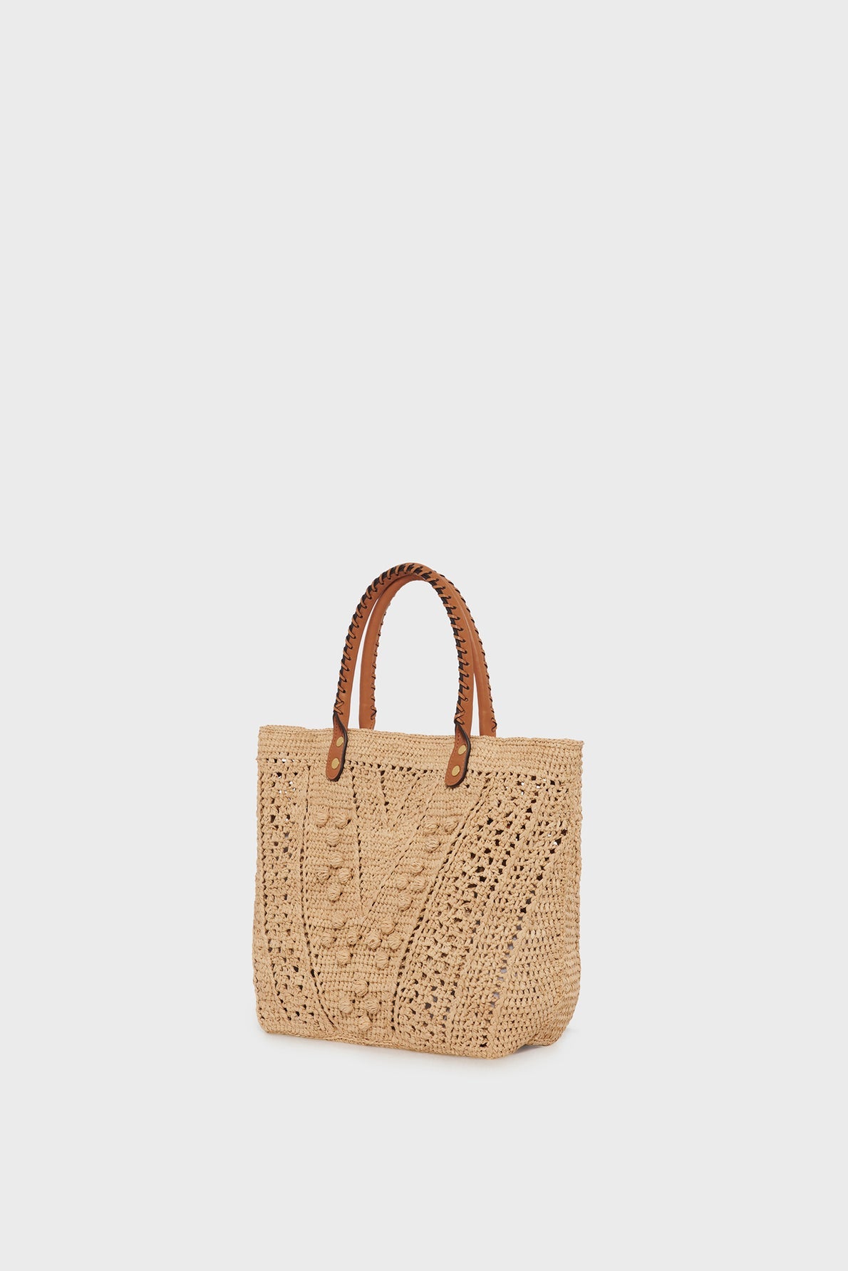 A beige crochet tote bag with brown leather handles is displayed against a plain white background. The bag, reminiscent of a sac à main raphia, features a textured, intricate woven pattern on the front that highlights its savoir-faire artisanal craftsmanship.