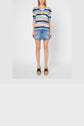 A woman stands wearing a blue, white, and yellow striped shirt with half sleeves, a denim mini skirt, and brown wedge sandals, against a plain white background.