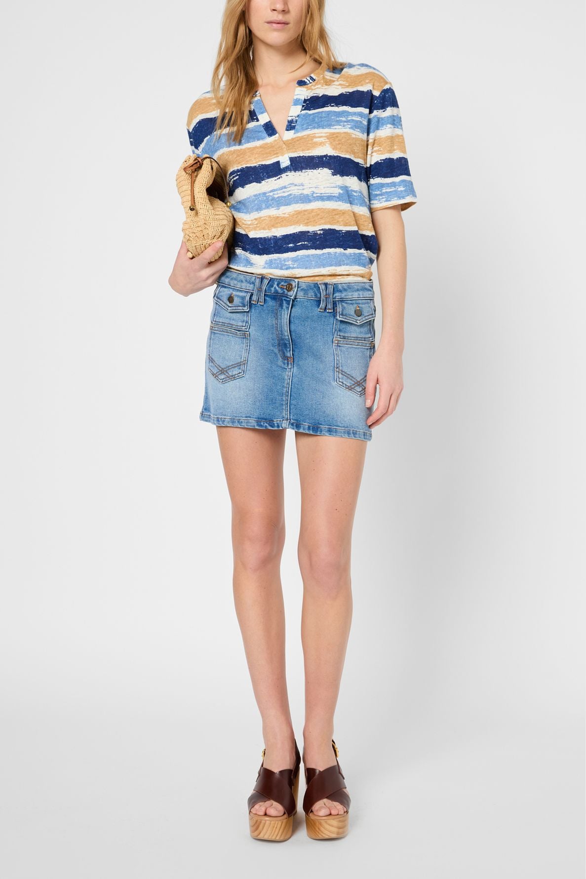 Woman wearing a blue, brown, and white striped shirt, denim mini skirt, and brown platform sandals, holding a woven clutch, standing against a plain light background.