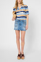 Woman wearing a blue, brown, and white striped shirt, denim mini skirt, and brown platform sandals, holding a woven clutch, standing against a plain light background.
