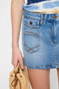 A person wearing a blue denim mini skirt holds a tan woven bag. The image shows the skirts front pocket details and part of the persons arm against a plain background.