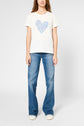 A woman stands facing forward, wearing a white t-shirt with a blue heart graphic, blue jeans, and white sneakers against a plain light background.
