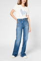A woman wearing a white t-shirt with a blue heart design, high-waisted blue jeans, and white sneakers stands smiling with one hand on her hip against a plain light background.