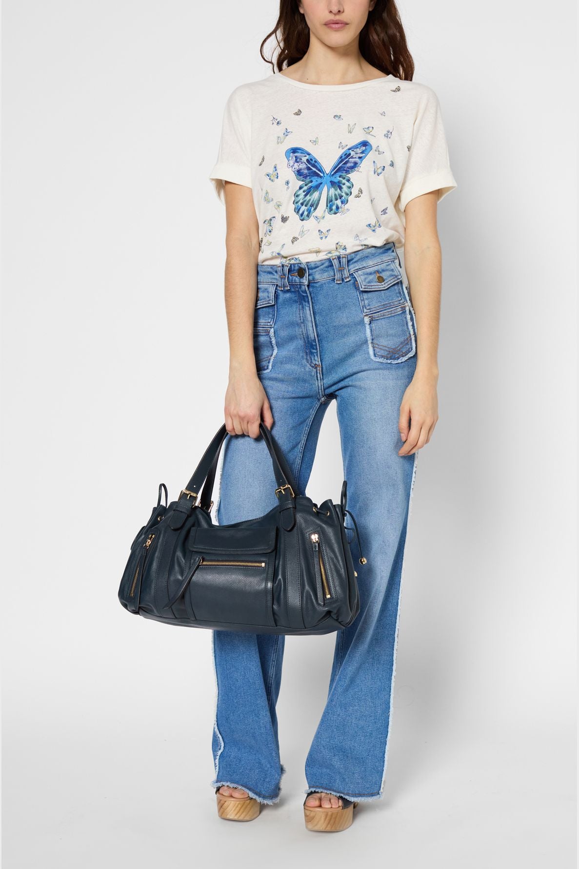 A woman wearing a white butterfly t-shirt, high-waisted blue jeans, and platform sandals holds a large black handbag by her side against a plain background.