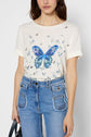 Woman wearing a white T-shirt with a large blue butterfly and small butterflies print, paired with high-waisted blue jeans with multiple pockets, holding a black handbag by her side.