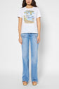 A woman stands facing forward, wearing a white graphic t-shirt with an illustration and text, light blue bootcut jeans with white side stripes, and beige platform sandals. The background is plain and light-colored.