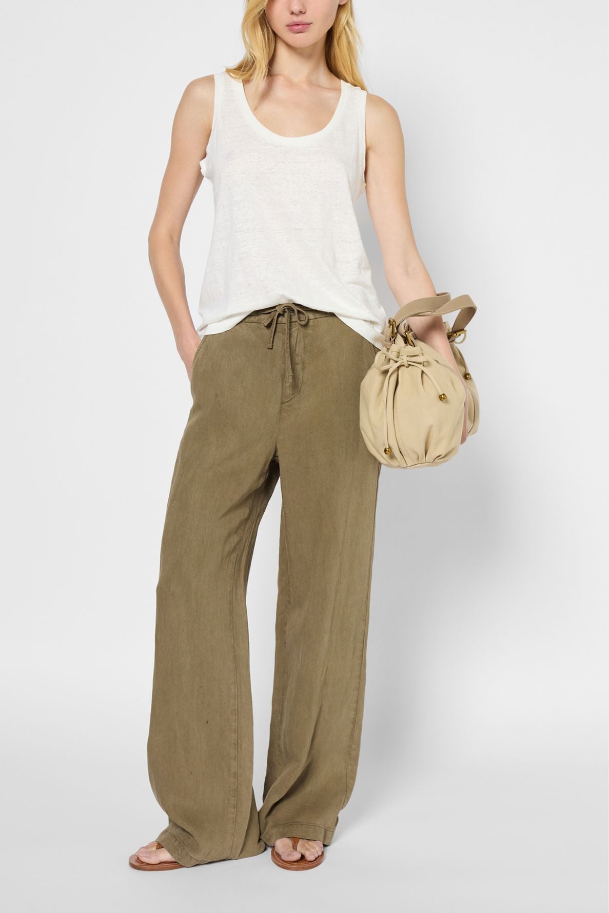 A woman wearing a débardeur lin blanc, loose olive green pants, and sandals holds a light beige round handbag. She stands against a plain light background.