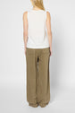 A person with long blonde hair is standing facing away, wearing a débardeur sans manche blanc in a breezy, sleeveless style perfect for summer, paired with loose-fitting, light brown pants. The background is plain and light-colored.