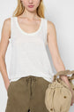 Woman wearing a white débardeur lin and olive green drawstring pants, holding a beige drawstring handbag. Only her upper body is visible against a plain background.