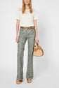 Woman wearing a white lace-detailed, manches courtes top, floral flared pants, brown belt, and wooden platform shoes, holding a wicker and leather handbag, standing against a plain background.