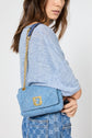 A woman in a light blue t-shirt ample casual and patterned jeans holds a small blue handbag with a gold chain strap and gold clasp, standing against a plain background.