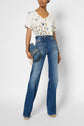A person wearing a white T-shirt oversize à col V with an imprimé papillons, high-waisted blue flared jeans, beige heels, and holding a blue woven clutch with a gold chain strap.