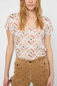 Woman wearing a short-sleeved patterned shirt with geometric designs and high-waisted brown pants with a hexagon texture, standing against a plain background. Her hair is loose and wavy.