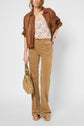 Woman wearing a brown leather jacket, patterned top, high-waisted tan pants, and platform sandals, holding a tan handbag. She stands against a plain white background.