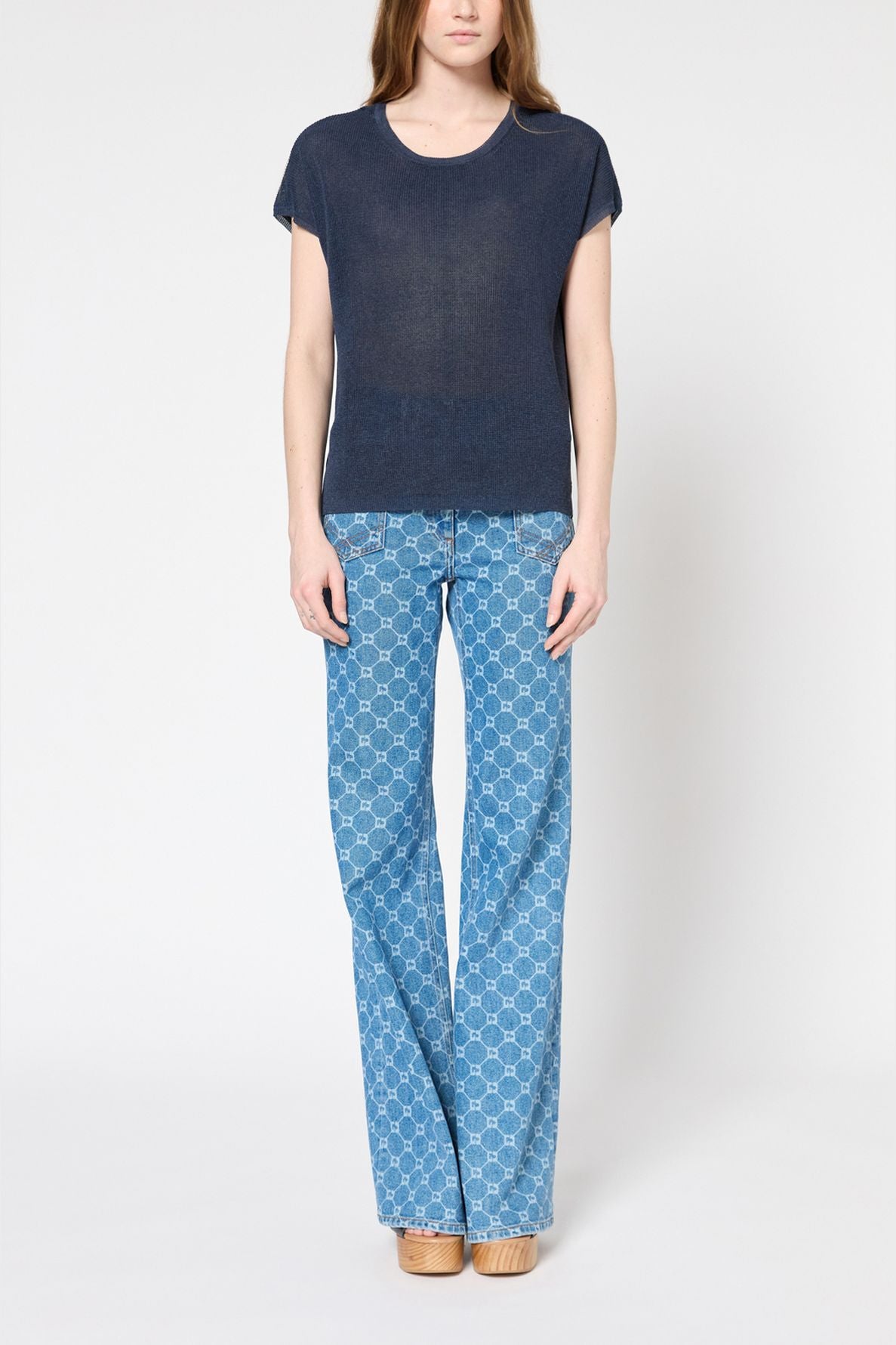 A person stands against a plain background, wearing a short-sleeve navy pull col rond, light blue patterned jeans, and beige platform shoes. Their straight, light brown hair falls over their shoulders.