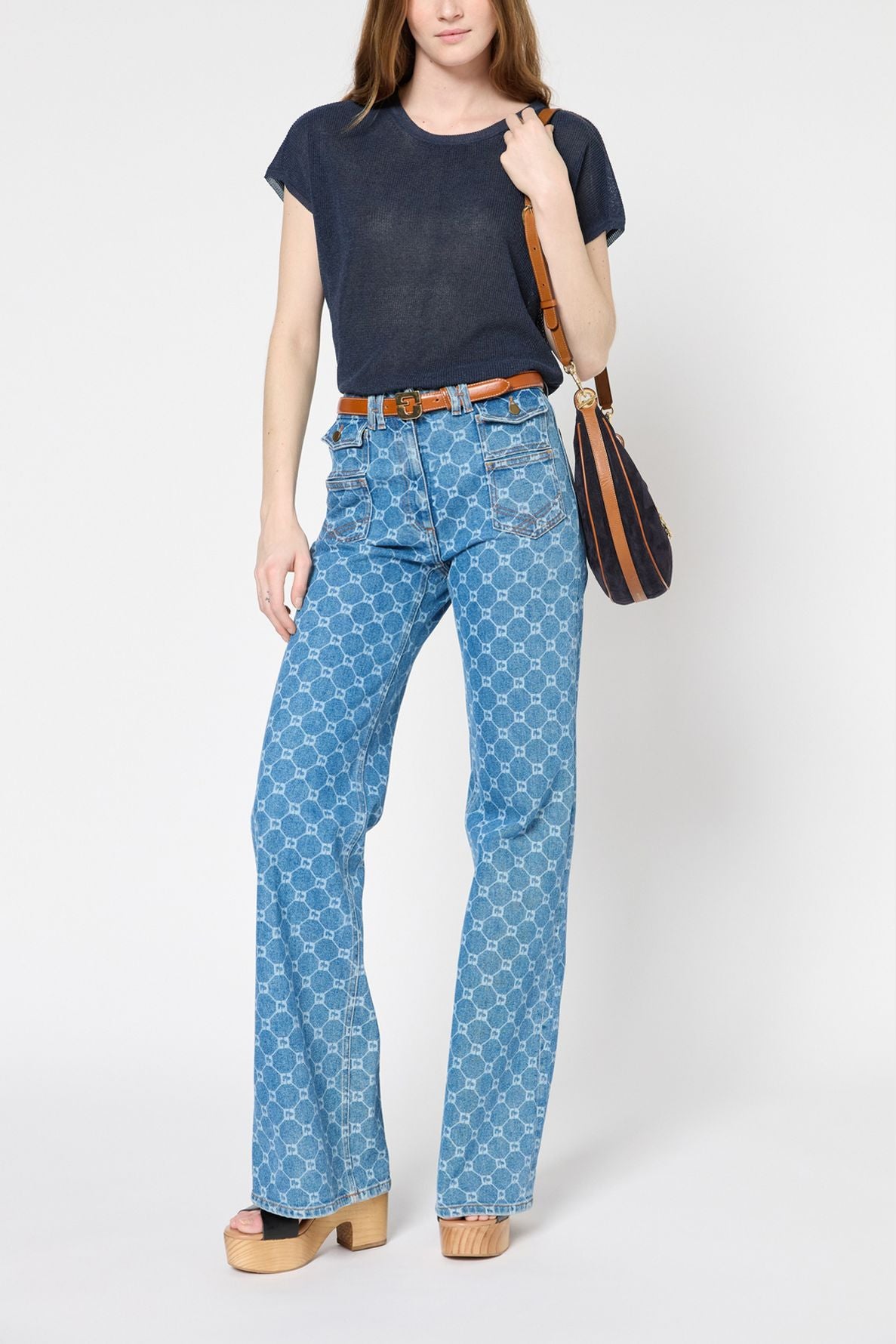 A woman wearing a navy blue pull col rond, patterned high-waisted flared jeans, tan platform sandals, a brown belt, and carrying a black and brown shoulder bag stands against a plain white background. The look is effortlessly stylish with elements that evoke the chic appeal of mode fabriqué en Italie.