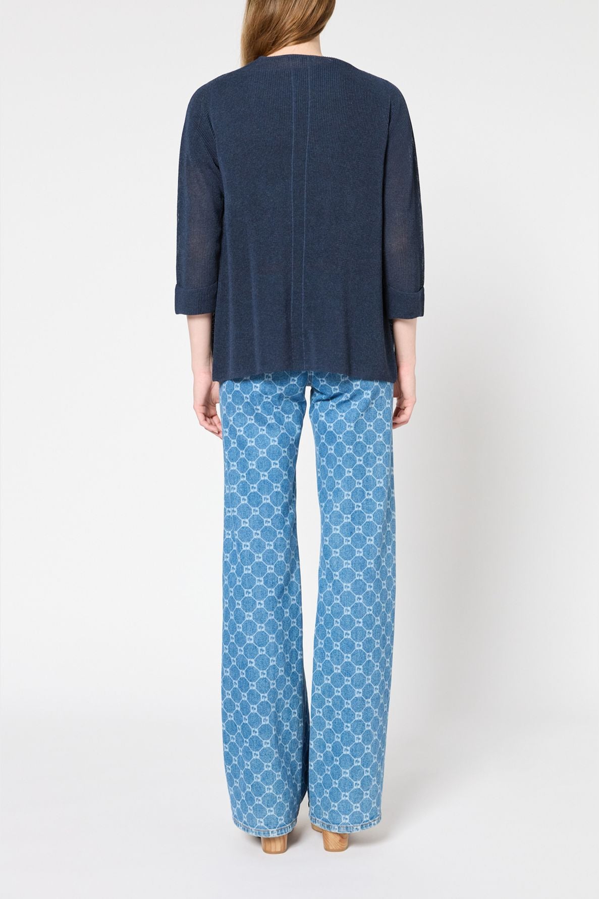 A person stands facing away, wearing a navy blue long-sleeve top and light blue patterned wide-leg jeans with beige platform shoes, against a plain white background.