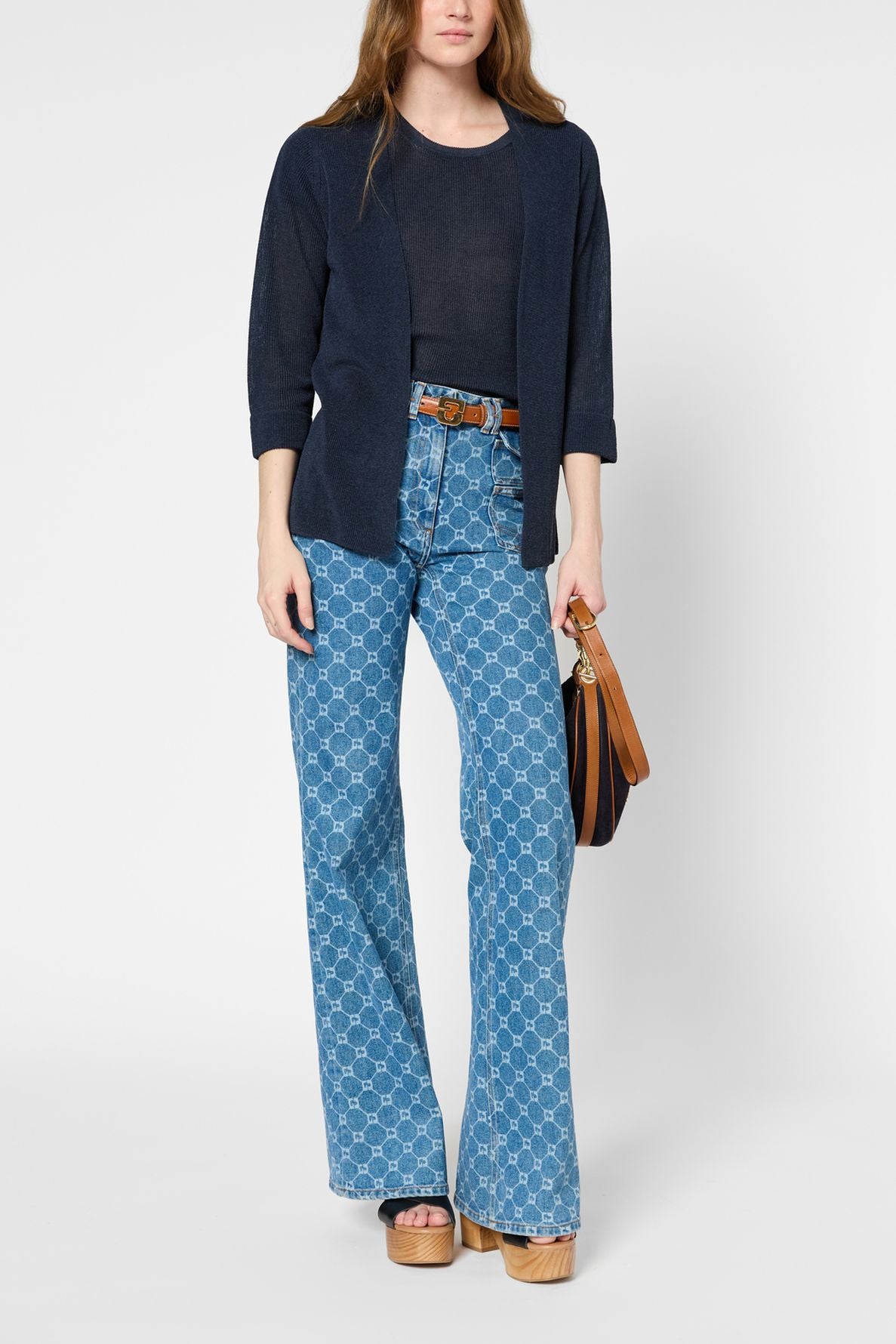 A person wearing a navy cardigan, black top, patterned high-waisted flared jeans, platform sandals, and holding a brown handbag.