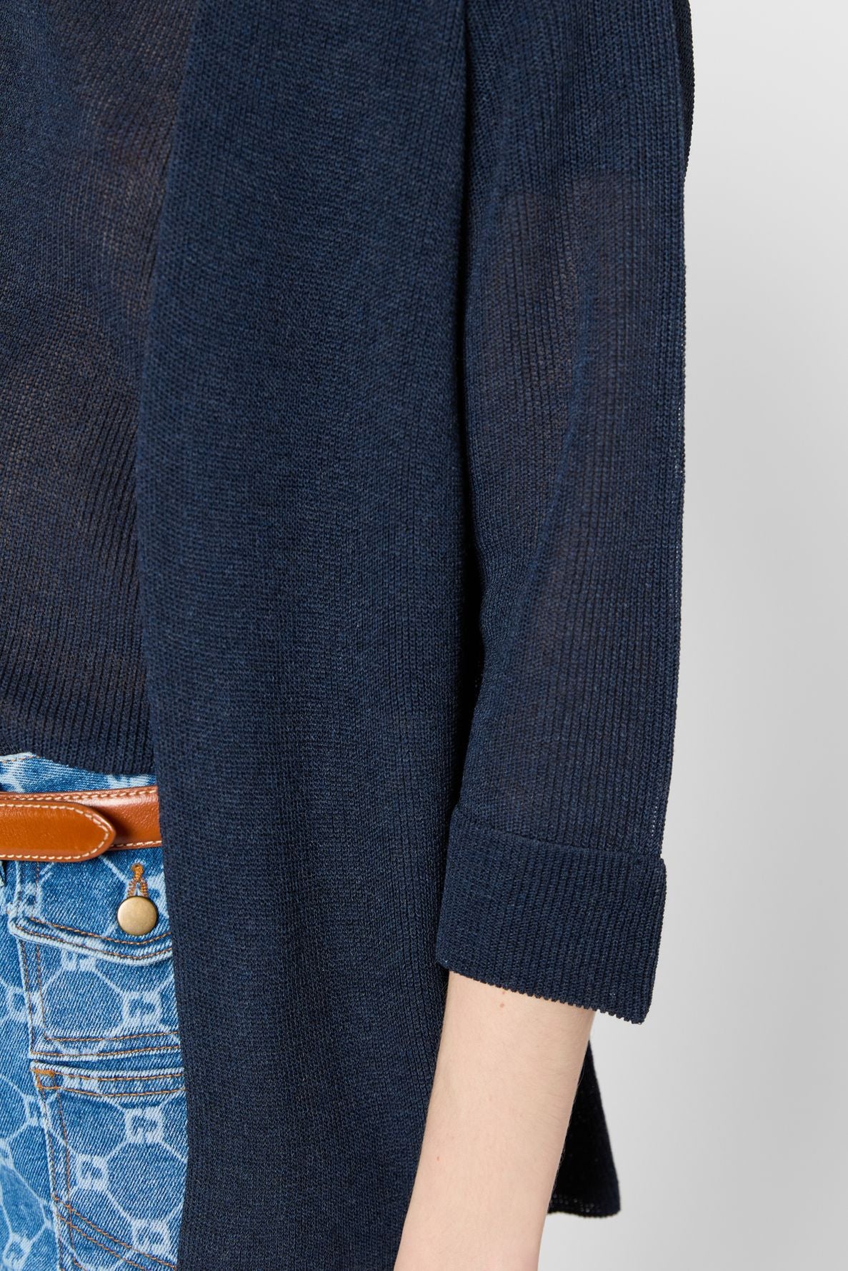 A person wearing a navy blue, long-sleeve, ribbed cardigan with rolled cuffs over a sheer top, and blue patterned jeans with a brown leather belt. Only the torso and arm are visible.