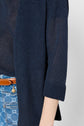A person wearing a navy blue, long-sleeve, ribbed cardigan with rolled cuffs over a sheer top, and blue patterned jeans with a brown leather belt. Only the torso and arm are visible.
