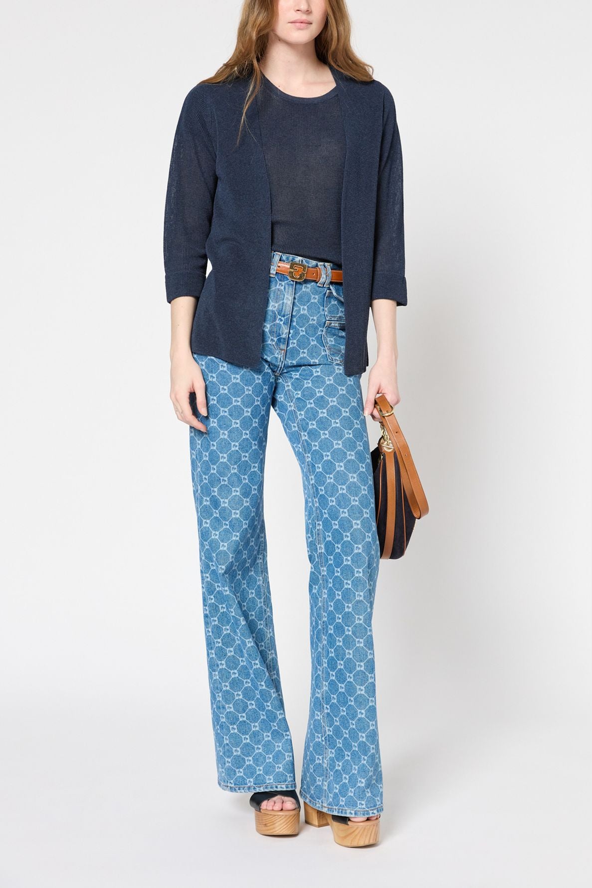 Woman standing against a white background wearing a navy cardigan, sheer navy top, patterned high-waisted wide-leg jeans, brown belt, platform sandals, and holding a brown handbag.