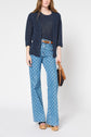 Woman standing against a white background wearing a navy cardigan, sheer navy top, patterned high-waisted wide-leg jeans, brown belt, platform sandals, and holding a brown handbag.