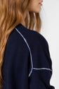 A woman with long, wavy brown hair is shown in profile, wearing a navy blue sweater with light-colored seam details. The background is plain and light-colored.