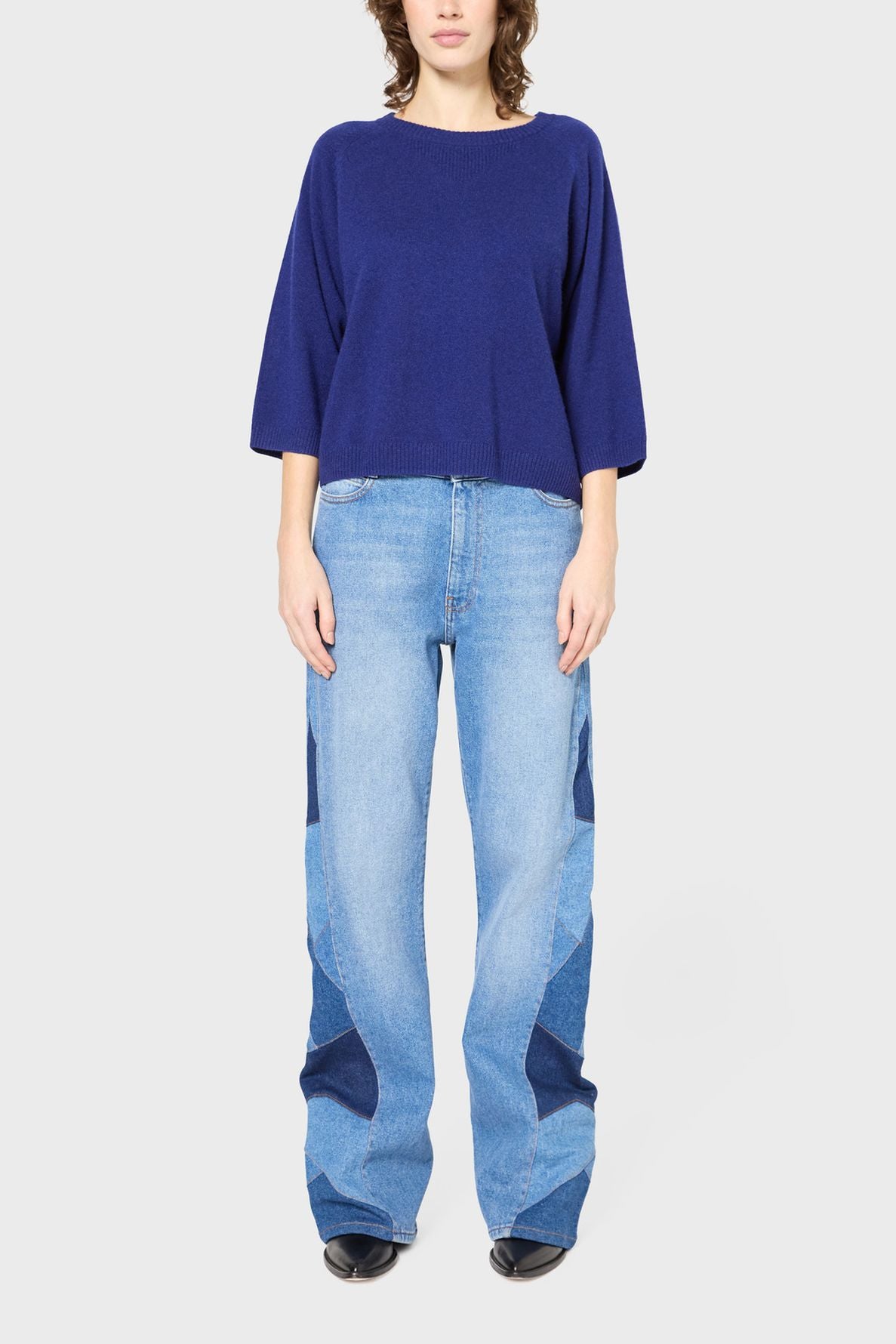 A person wearing a loose, blue sweater and wide-leg, patchwork light blue jeans stands facing forward against a plain white background.